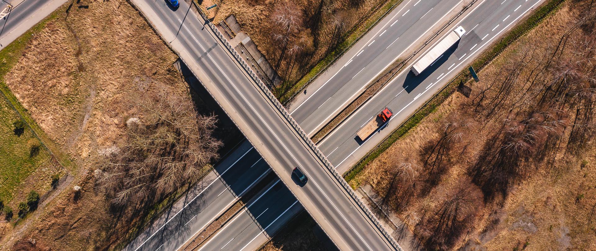 Obraz przedstawia widok z lotu ptaka na skrzyżowanie dróg z wiaduktem. Wiadukt biegnie ukośnie od lewego dolnego rogu do prawego górnego, przecinając prostopadłą drogę poniżej. Na obu drogach widoczne są liczne pojazdy, w tym samochody i ciężarówka. Okoliczny teren jest trawiasty, z kilkoma drzewami i fragmentami gołej ziemi. 