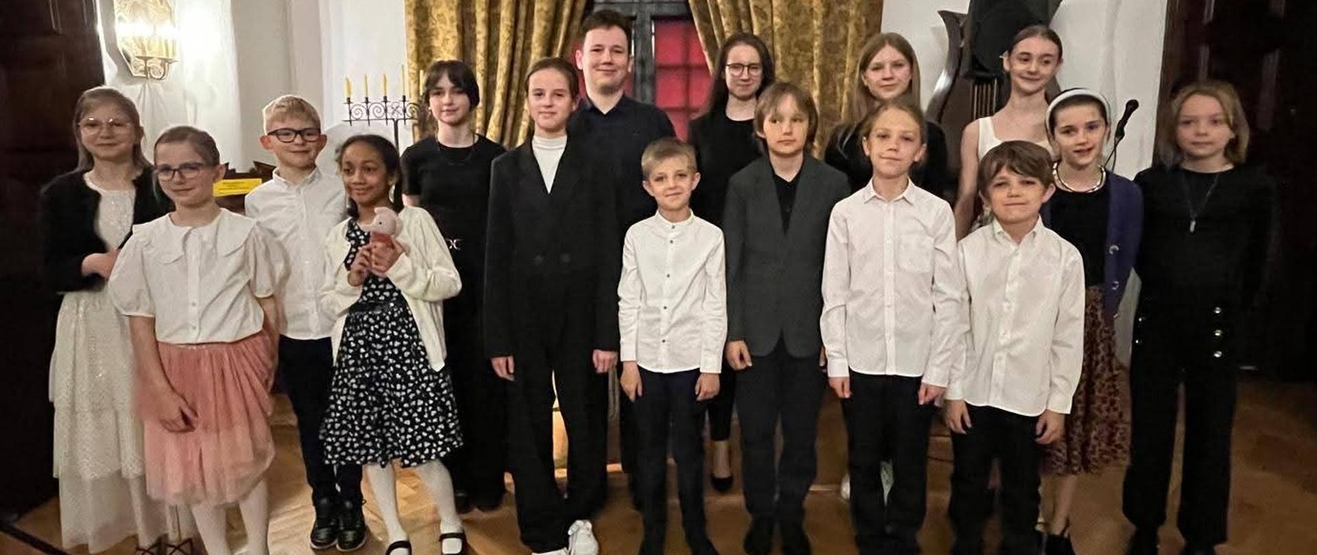Zdjęcie kolorowe, grupa piętnastu dzieci i jedna dorosła osoba stoi w eleganckim wnętrzu z parkietem i złotymi zasłonami w tle. Dzieci ubrane są odświętnie – chłopcy w białych koszulach, marynarkach lub garniturach, dziewczynki w sukienkach, spódnicach i kardiganach. Niektóre dzieci noszą okulary. W tle widoczne są świecznik, instrument muzyczny oraz ozdobne oświetlenie na ścianie. Wszyscy patrzą w stronę aparatu i uśmiechają się.