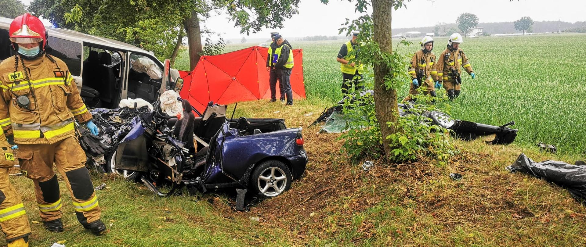 Na zdjęciu rozbity samochód osobowy w rowie koloru granatowego oraz bus również w rowie obok którego stoją policjanci, na zdjęciu również strażacy podczas działań 