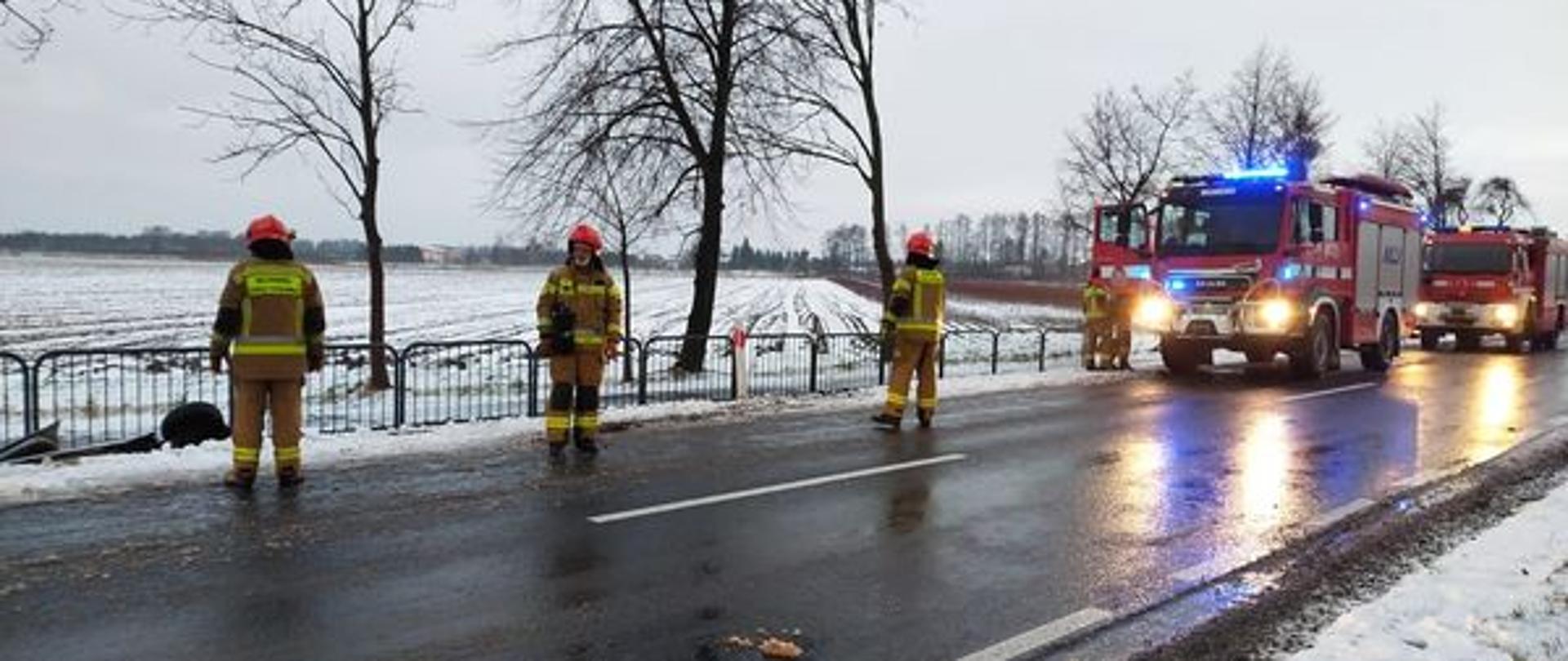 Zima, biało od śniegu na poboczu drogi. Na drodze z prawej strony stoją dwa samochody strażackie na światłach, na drodze stoi trzech strażaków w jasnych mundurach i jasnych hełmach.