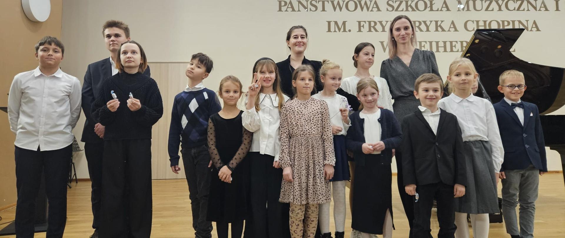 Na sali koncertowej pozują na stojąco do zdjęcia uczniowie klasy fortepianu Julianny Bartyzel oraz Katarzyny Dzierzgowskiej.