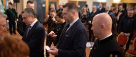 Minister Czarnek i stojący obok starszy mężczyzna w garniturze rozmawiają z ludźmi w czarnych galowych mundurach, minister trzyma w ręku otwartą małą książeczkę, obok stoi mężczyzna w czarnym galowym mundurze, dalej sala z żółtymi ścianami wypełniona ludźmi.
