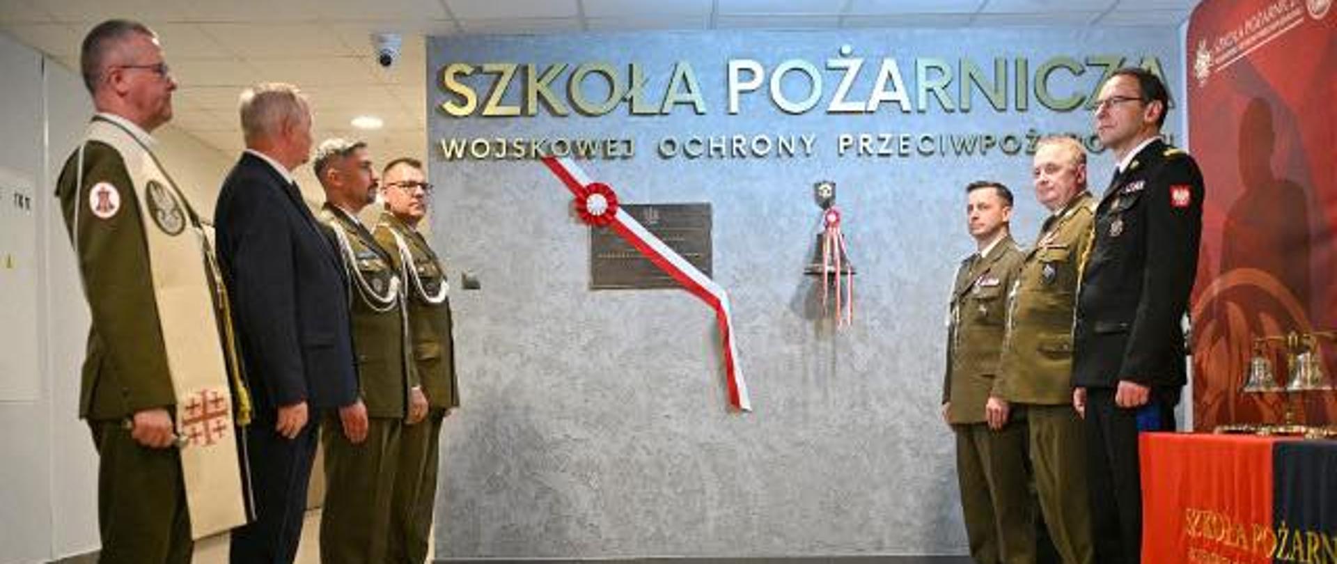 Uroczystość odsłonięcia tablicy pamiątkowej w Szkole Pożarniczej Wojskowej Ochrony Przeciwpożarowej. Przed ścianą z nazwą szkoły stoją funkcjonariusze i przedstawiciele kierownictwa w mundurach oraz garniturach. Obok tablicy znajdują się biało-czerwone wstęgi.