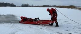 Dwóch strażaków znajduje się na zamarzniętej tafli wody. Jeden ze strażaków leży na brzuchu na deskosaniach. Drugi strażak trzymając tylne uchwyty na deskosaniach przemieszcza się po zamarzniętym zbiorniku wodnym.
Strażacy ubrani w kombinezony wypornościowe koloru czerwonego, zabezpieczeni linkami ratowniczymi. Tafla lodu pokryta śniegiem, w tle widoczne drzewa.
