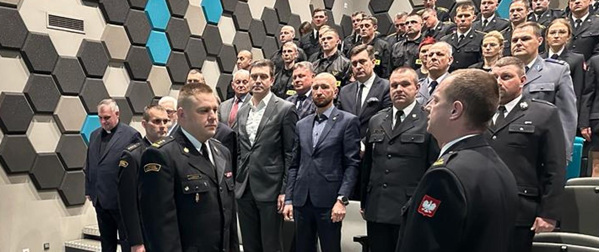 składanie muldunku przez strażaka drugiemu strażakowi, w tle sala i duża grupa osób, wszyscy stoją na baczność