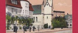 Przedwojenna pocztówka, wyblakłe kolory, przedstawiająca budynek szkoły muzycznej wraz z sąsiadującym obiektem kościoła. Kościół strzelisty, biały z ciemną dachówką wierzy, budynek szkoły dwupiętrowy, biały z poprzecznym szarym pasem na wysokości pierwszego piętra i dachem mansardowym pokrytym czerwoną dachówką. Przed budynkiem rosną drzewa, posesja odgrodzona od ulicy na której widać przechodniów.