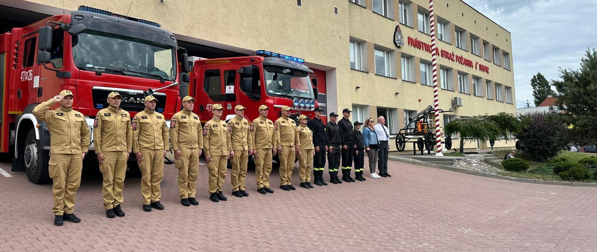 Na zdjęciu widać strażaków w szyku w żółtych oraz czarnych ubraniach służbowych. W szyku stoją również dwie osoby kobieta oraz mężczyzna w ubraniach cywilnych. Za szykiem stoją dwa pojazdy gaśnicze. Widać flagę na szacie.