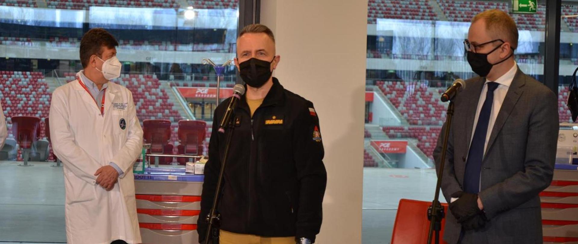 Zdjęcie przedstawia Komendanta Głównego PSP nadbryg. Andrzeja Bartkowiaka na briefingu prasowym w szpitalu tymczasowym na PGE Narodowy. Komendant w czarnej bluzie polarowej i maseczce stoi przed mikrofonem. Po lewej stronie zdjęcia znajduje się lekarz w białym fartuch i maseczce, po prawej wiceminister MSWiA Błażej Pobożny w garniturze koloru szarego i maseczce. Za placami komendanta przez oszklenie widoczne są sektory stadionu