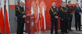 Strażacy stoją na tle ścianki firmowej Straży Pożarnej, Komendant przemawia z mównicy, jeden z gości trzyma kwiaty.