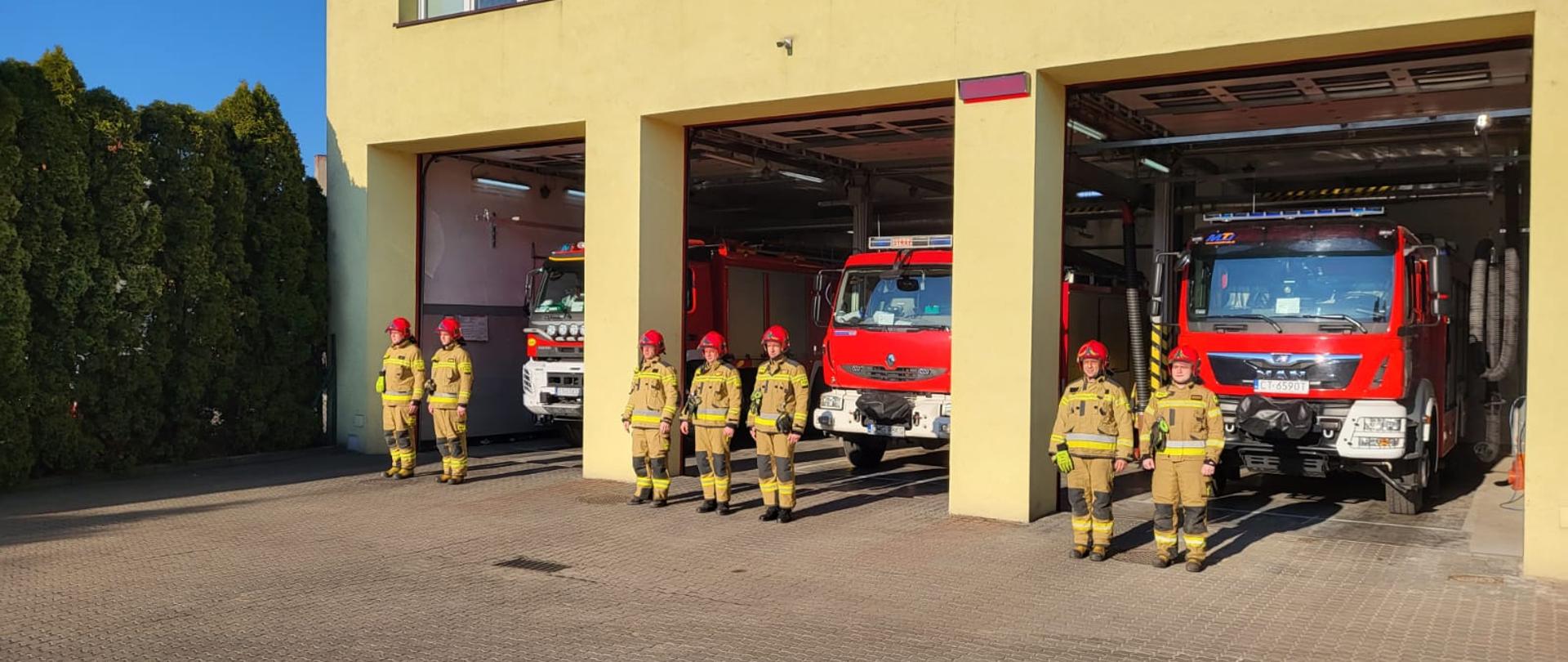 Strażacy stojący przed garażem oddają hołd poległym strażakom z Ukrainy. Otwarte bramy garażowe. W garażu 3 pojazdy pożarnicze.