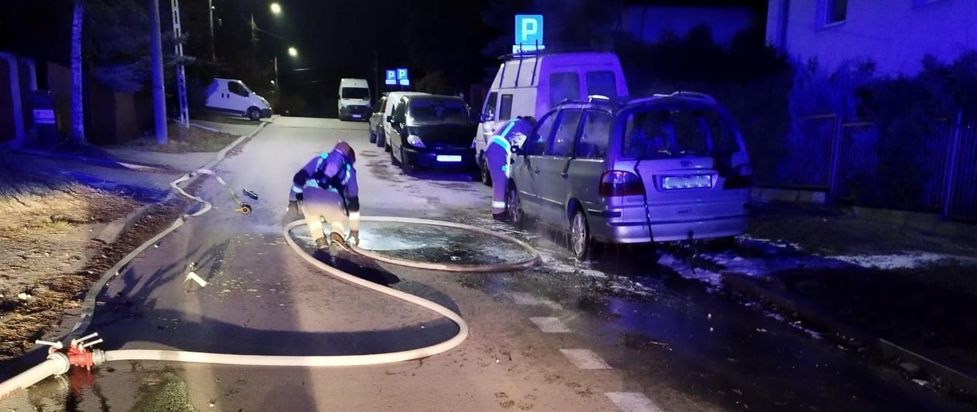 Zdjęcie przedstawia ulicę, po prawej stronie znajduje się zniszczony przez pożar samochód osobowy. Na jezdni rozciągnięte leżą węże gaśnicze przy których pracuje ratownik. Drugi ratownik prowadzi działania przy spalonym samochodzie. 