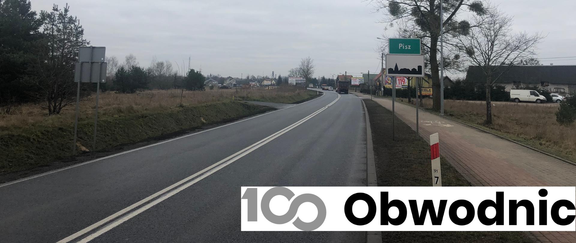 Droga asfaltowa, po prawej stronie ścieżka rowerowa oraz znak informacyjny z nazwą miasta i oznaczeniem obszaru zabudowanego. W tle zabudowania.