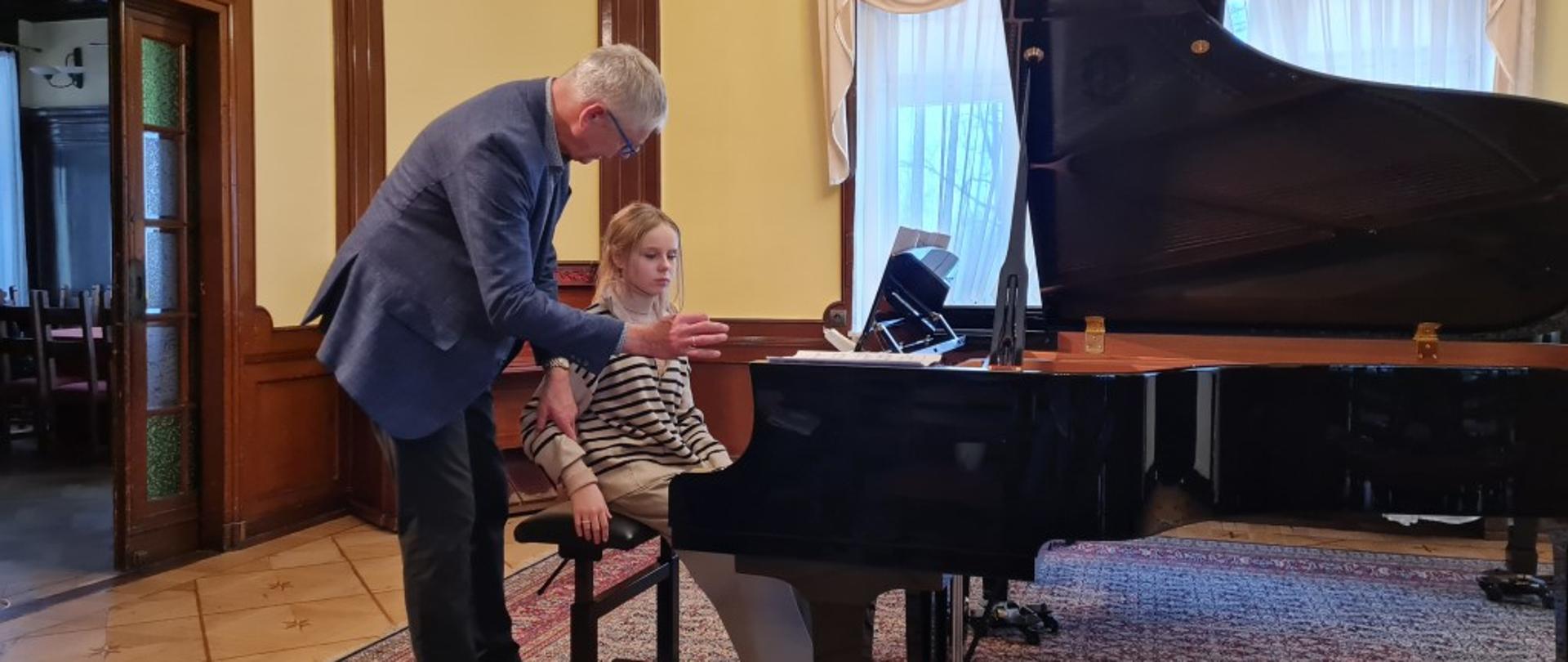 Zdjęcie kolorowe. Dziewczynka z długimi blond włosami, w beżowym sweterku w paski siedzi na taborecie przy fortepianie, za nią stoi siwy mężczyzna w okularach i granatowej marynarce, które pokazuje prawą rękę