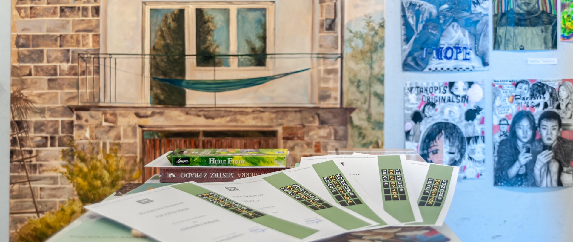 Na zdjęciu widać stół, na którym leżą dyplomy, w tle znajduje się duży obraz przedstawiający balkon z zielonym hamakiem oraz kilka kolorowych prac plastycznych powieszonych na ścianie.