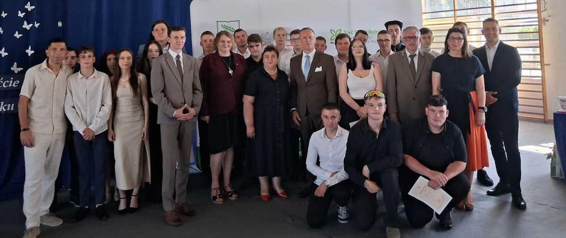 Zdjęcie przedstawia dużą grupę ludzi pozujących do wspólnej fotografii wewnątrz hali sportowej lub sali gimnastycznej.Otoczenie i tłoSufit: Wysoki, o konstrukcji z drewnianych belek i metalowych rur.Tło: Za ludźmi wisi duża, ciemnoniebieska kurtyna (po lewej ozdobiona białymi motylkami).Środek tła: Biała ścianka wystawowa z logo i napisami (widoczne m.in. „Sieć Rozwoju”).Prawa strona: Kosz do koszykówki oraz duże okna wpuszczające dzienne światło.Ludzie na zdjęciuWydarzenie ma charakter oficjalny, na co wskazuje elegancki ubiór większości zebranych. Grupa składa się z około 25 osób – młodzieży oraz dorosłych.Pierwszy plan (osoby kucające/siedzące): Trzech młodych mężczyzn z przodu. Ten po prawej trzyma w dłoni biały dyplom lub certyfikat.Główna grupa (stojący w centrum):Kobiety w eleganckich sukienkach (czarnych, kremowych) oraz ciemnych kompletach.Mężczyźni w garniturach (brązowym, szarym, czarnym), koszulach oraz jeden w mundurze leśnika (zielony garnitur).Lewa strona: Grupa młodzieży, w tym chłopak w białej koszuli i dziewczyna w beżowej sukience. Na skraju po lewej stoi mężczyzna w jasnym, luźniejszym komplecie.Wszyscy są zwróceni przodem do obiektywu, większość osób delikatnie się.