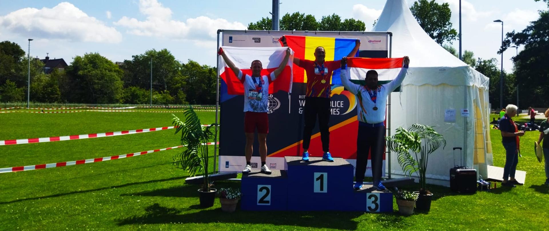 Trzech zawodników stoi na podium, maja uniesione flagi za plecami.