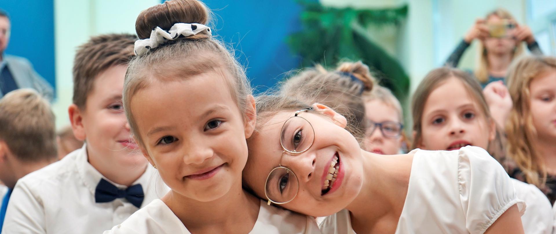 Na sali siedzą dzieci, z przodu dwie uśmiechnięte dziewczynki w białych sukienkach.