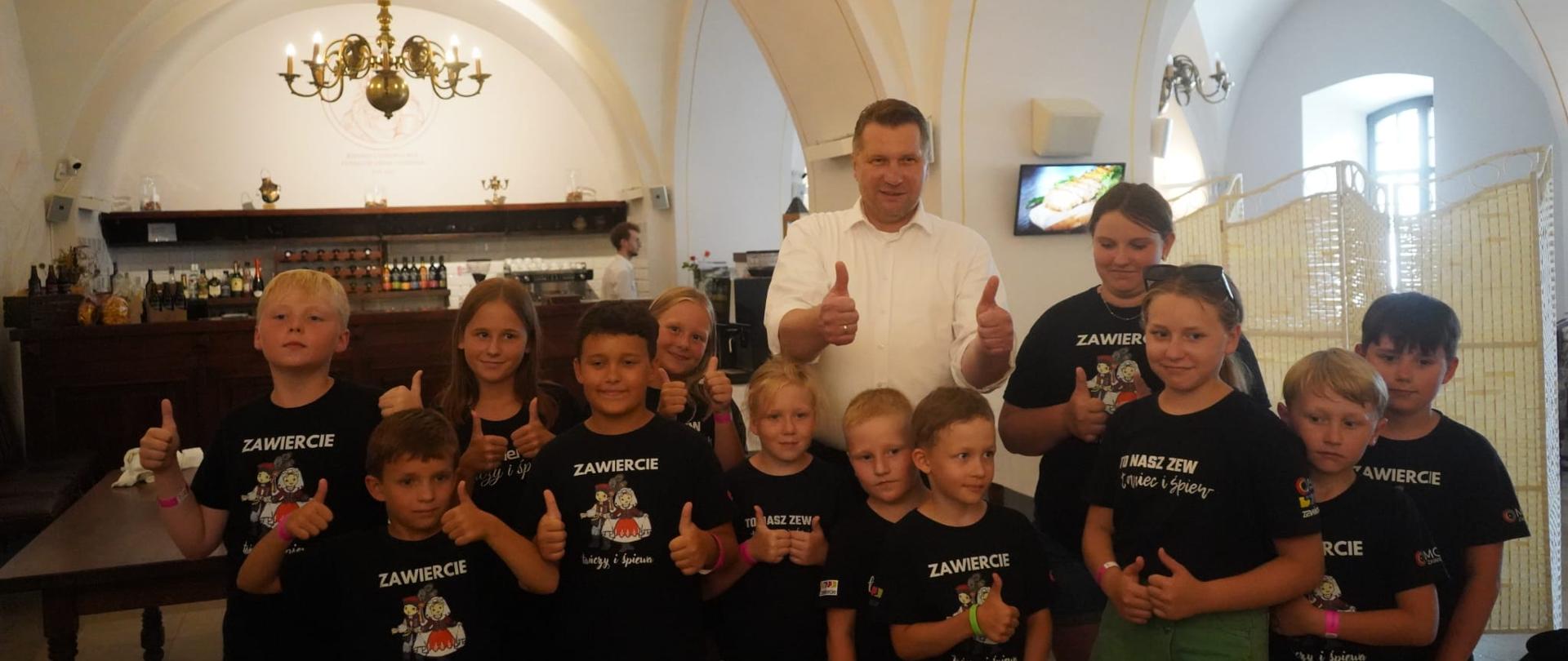 Grupa dzieci z mężczyzną pozuje do zdjęcia wyciągając kciuki w geście ok. Dzieci ubrane są w koszulki z napisem Zawiercie.