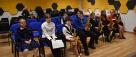 Na zdjęciu widnieje grupa uczniów siedzących w sali kameralnej PSM I stopnia w Jaśle. Niektórzy z uczniów trzymają w rękach saksofony. Przeważająca kolorystyka zdjęcia: brązowo-czarno-niebiesko-żółta.
