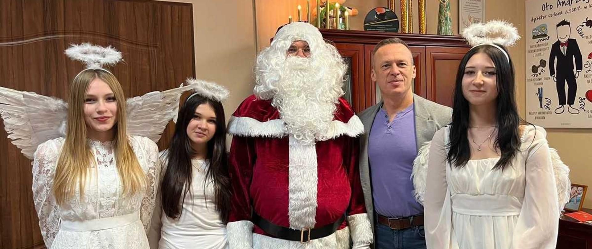 Zdjęcie przedstawia pięć osób, w tym cztery młode kobiety i jednego mężczyznę w średnim wieku, pozujące razem w świątecznej atmosferze, prawdopodobnie w biurze lub podobnym pomieszczeniu.
Centralna postać: W centrum kadru stoi mężczyzna przebrany za Świętego Mikołaja, w tradycyjnym, bordowym stroju z białymi futrzanymi wykończeniami i szerokim czarnym pasem. Ma na sobie dużą, białą brodę i wąsy.
Anioły: Po lewej i prawej stronie Mikołaja stoją cztery młode kobiety ubrane w białe sukienki (dwie w długie, koronkowe sukienki po lewej stronie, jedna w krótką, gładką sukienkę obok Mikołaja, i jedna w długą, luźną sukienkę po prawej). Wszystkie mają białe skrzydła i aureole na głowach, stylizowane na anioły.
Mężczyzna: Między Mikołajem a kobietą po prawej stronie stoi drugi mężczyzna, ubrany w fioletową koszulkę polo, szarą marynarkę i dżinsy.
Tło: W tle znajduje się drewniana szafka z różnymi dekoracjami, w tym z trofeami i świecami, oraz plakaty na ścianie. Jeden z plakatów ma napis ""Oto Andrzej"". Oświetlenie jest ciepłe i świąteczne.