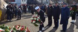 Żołnierz Marynarki Wojennej RP składa wiązankę pod pomnikiem za nim stoi delegacja służb mundurowych służb podległych Ministerstwu Spraw Wewnętrznych i Administracji obok stoją zaproszeni goście.