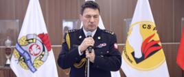 Odchodzący na emeryturę Komendant Centralnej Szkoły PSP przemawiający do mikrofonu podczas uroczystego apelu, w tle flagi z logo PSP oraz CS PSP