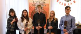 Zdjęcie przedstawia uczniów z klasy gitary p. Anny Pietrzak wraz z nauczycielką. Na scenie stoją: p. Anna Pietrzak, Weronika Malinowska, Jan Berłowski, Karolina Kuca i Michał Ziernik. Za uczniami stoi czarny fortepian, w tle znajdują się okna a na firanach zawieszone są świąteczne ozdoby i plakat informujący o konkursie.