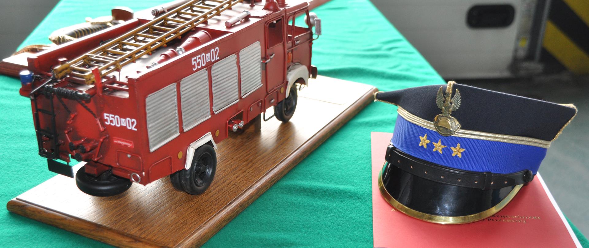 Fotografia przedstawia replikę pojazdu gaśniczego marki Star 244 z naniesionymi numerami operacyjnymi zastępcy komendanta "550-02". Obok pojazdu czapka rogatywka do ubioru galowego.