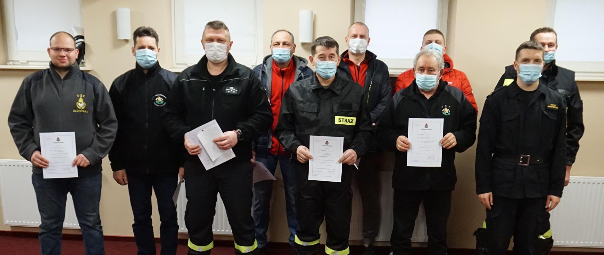 W pomieszczeniu na czerwonym dywanie stoją w dwóch rzędach strażacy w czarnych mundurach oraz ubraniach cywilnych.