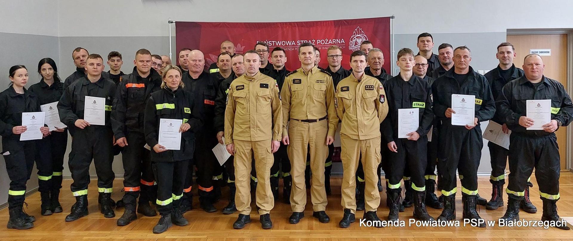 28 strażaków ochotniczej straży pożarnej w umundurowaniu czarnym koszarowym oraz w środku trzech funkcjonariuszy państwowej straży pożarnej w umundurowaniu służbowym w kolorze piaskowym. W tle czerwony baner państwowa straż pożarna. Strażacy ochotnicy trzymają zaświadczenia o ukończeniu szkolenia podstawowego.
