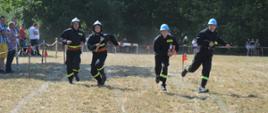 Zawody sportowo-pożarnicze w Jeziorach. Plac ćwiczeń. Trwa jedna z konkurencji zawodów - sztafeta pożarnicza 7x50 m z przeszkodami. czterech zawodników biegnie po torze. W tle drzewa, przeszkody i kibice.