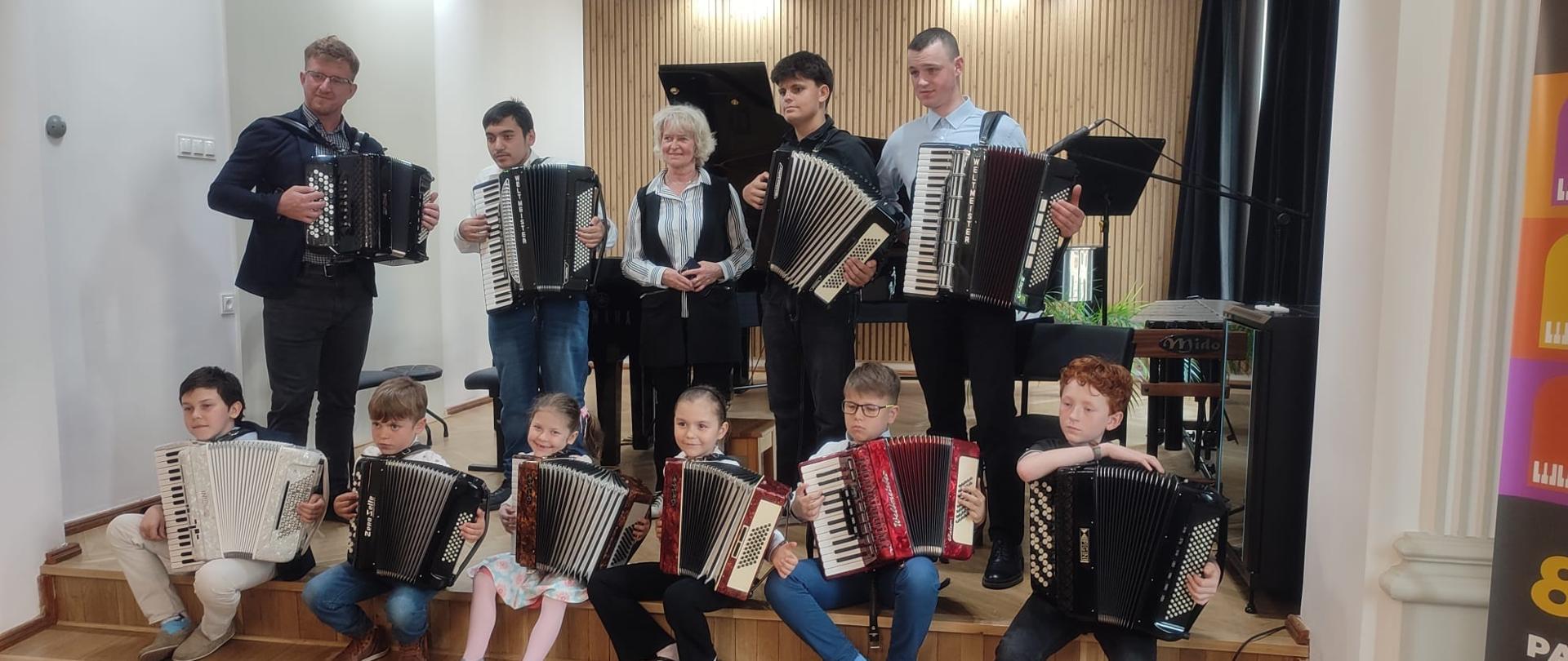 Galeria 11 zdjęć z koncertu klasy akordeonu uczniów p. Liliany Stopki i p. T. Brusińskiego, który odbył się 30 maja 2025 w auli PSM w Kłodzku. Uczniowie grają na akordeonie w sali koncertowej.