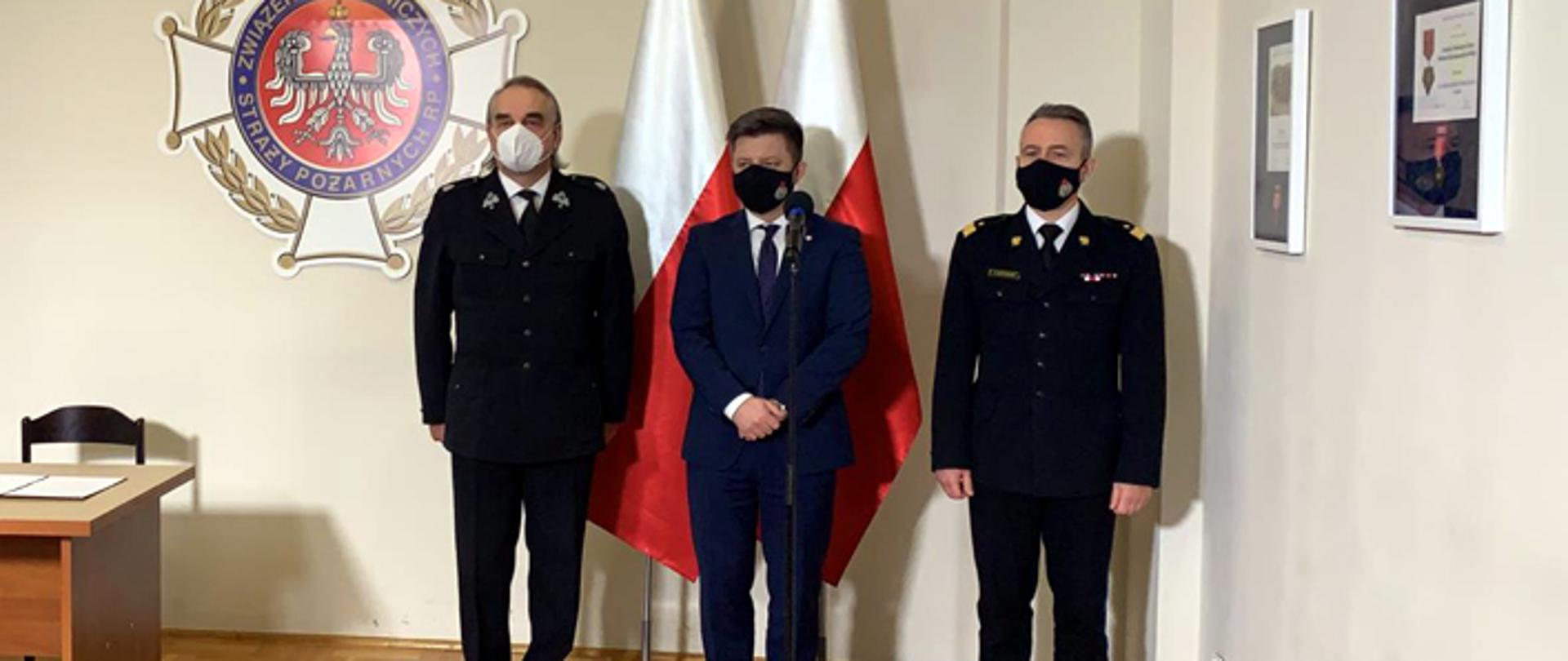 Zdjęcie przedstawia stojących na tle biało czerwonych flag kolejno od lewej dh. Waldemara Pawlaka Prezesa ZG ZOSP RP, Michała Dworczyka, szefa Kancelarii Prezesa Rady Ministrów oraz nadbryg. Andrzeja Bartkowiaka Komendanta Głównego PSP.