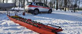 Zbliżenie na pomarańczowe sanie lodowe leżące na śniegu. Na saniach umieszczone jest wiosło i żółty worek z linką. W tle stoi srebrny samochód terenowy z czerwonym pasem i napisem "STRAŻ" oraz numerami operacyjnymi. Obok samochodu widać przyczepę z łodzią. Całość znajduje się na zaśnieżonym terenie w pobliżu parku z wysokimi, suchymi drzewami.