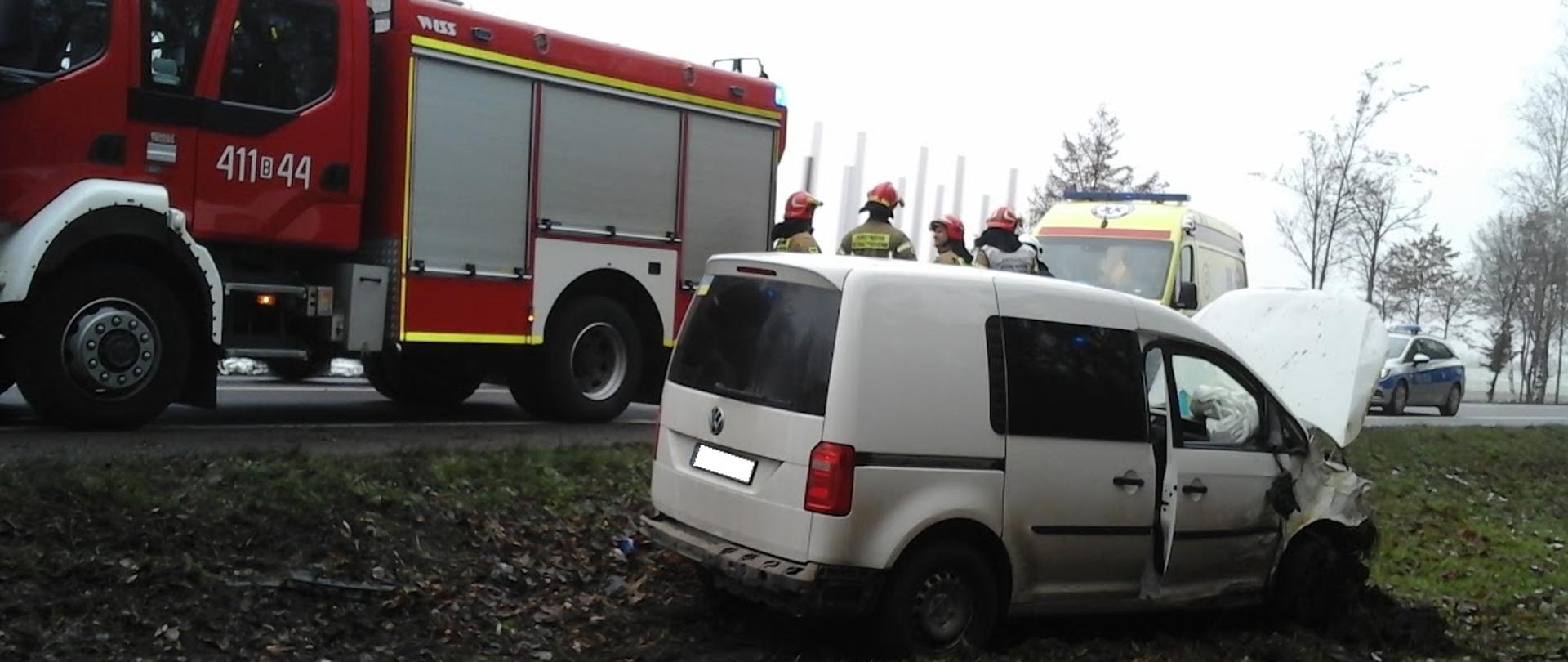 Samochód osobowy po wypadku stoi na poboczu drogi. Auto ma uszkodzony przód. W tle samochód strażacki, karetka i radiowóz stojące na jezdni.