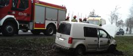 Samochód osobowy po wypadku stoi na poboczu drogi. Auto ma uszkodzony przód. W tle samochód strażacki, karetka i radiowóz stojące na jezdni.