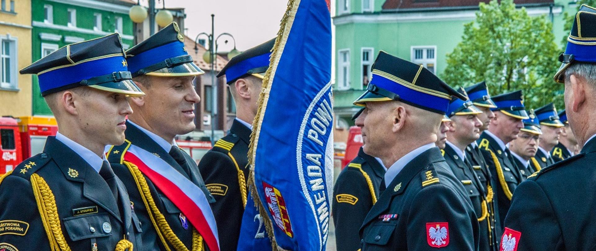 strażak ściska rękę drugiemu strażakowi, który ma na sobie biało czerwoną szarfe oraz trzyma sztandar