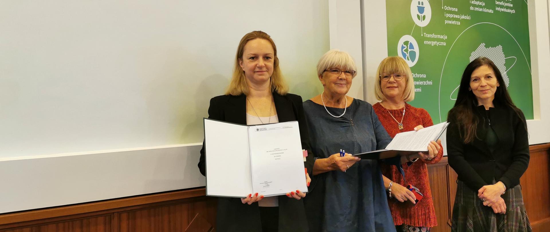 Zdjęcie przedstawia moment po podpisaniu dofinansowania na przedsięwzięcie pod tytułem „Zmniejszenie zużycia energii i emisji CO2 z wykorzystaniem odnawialnych źródeł energii w 6 Zakładach Leczniczych Stowarzyszenia MONAR”. Do zdjęcia pozują (zaczynając od lewej strony) zastępca dyrektora w departamencie transformacji energetycznej w Narodowym Funduszu Ochrony Środowiska i Gospodarki Wodnej Magdalena Bodzenta, przewodnicząca zarządu głównego stowarzyszenia MONAR Elżbieta Zielińska, sekretarz zarządu głównego stowarzyszenia MONAR Jagoda Władoń- Wilecka oraz koordynator projektu ze strony Narodowego Funduszu Ochrony Środowiska i Gospodarki Wodnej Joanna Jaworek. Panie stoją na tle loga Narodowego Funduszu Ochrony Środowiska i Gospodarki Wodnej i trzymają w rękach teczki z podpisanymi umowami.