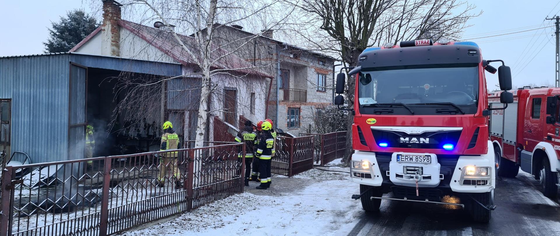 Na drodze asfaltowej przy brązowym ogrodzeniu zabudowy jednorodzinnej stoją dwa samochody strażackie. Na lewo od samochodów, w bramie ogrodzenia stoi 3 strażaków w granatowych strojach bojowych i skierowani są twarzą do blaszanego garażu, w którym znajdują się 2 strażacy w piaskowych strojach bojowych. Za blaszanym garażem znajdują się dwa domy jednorodzinne.