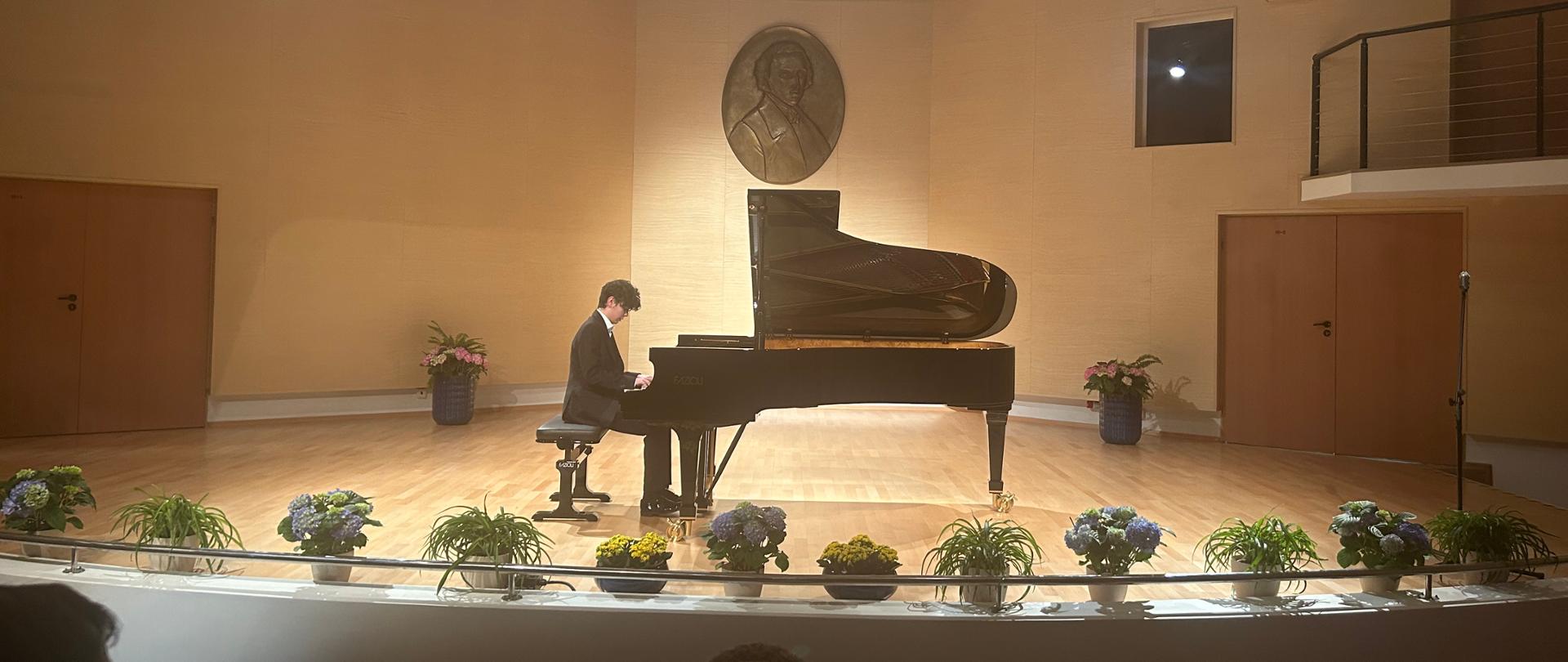 Na scenie sali koncertowej znajduje się fortepian marki Fazioli, wokół sceny znajdują się kolorowe kwiaty w doniczkach. Przy fortepianie siedzi artysta grający utwór