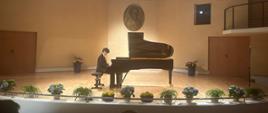 Na scenie sali koncertowej znajduje się fortepian marki Fazioli, wokół sceny znajdują się kolorowe kwiaty w doniczkach. Przy fortepianie siedzi artysta grający utwór