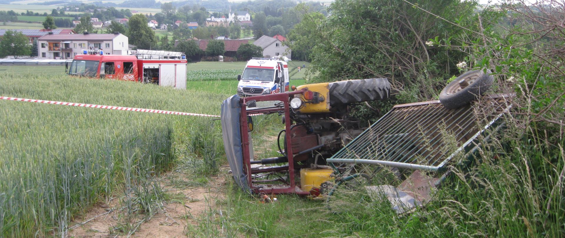 Na zdjęciu widoczny przewrócony rciągnik z zapietym wózkiem, w dali widoczy samochód pozarniczy oraz karetka ZRM