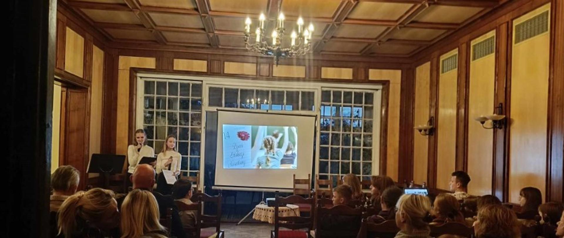 Zdjęcie kolorowe. Publiczność w sali kameralnej szkoły. Na scenie dwie nastolatki w strojach galowych. W tle ekran z ozdobnym napisem "Dzień Edukacji Narodowej" oraz białe okno. 