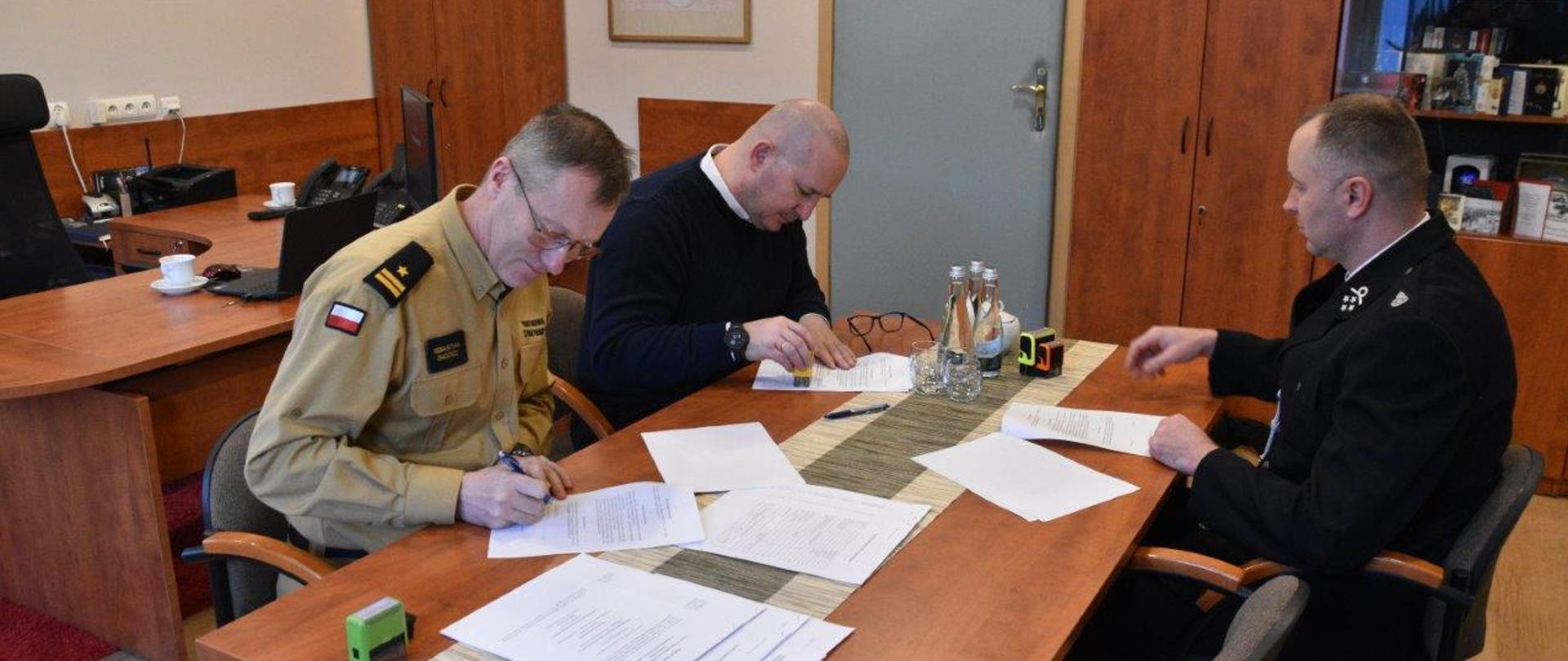 Na zdjęciu widnieje 3 mężczyzn. Podpisują oni porozumienie dotyczące włączenie jednostki Ochotniczej Straży Pożarnej w Potoku Wielkim w struktury krajowego systemu ratowniczo-gaśniczego. Pierwszy z lewej to Komendant Powiatowy PSP w Jędrzejowie, obok znajduje się Burmistrz Miasta i Gminy Jędrzejów, natomiast po przeciwnej stronie prezes tamtejszej jednostki w umundurowaniu wyjściowym.