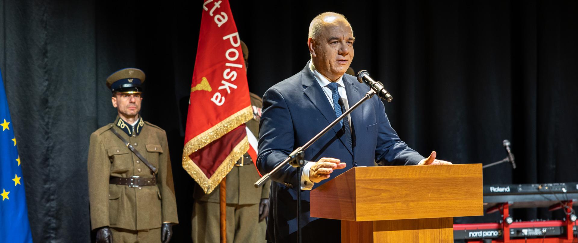 Wicepremier Jacek Sasin stoi za mównicą. W tle dwóch mężczyzn i sztandar. 