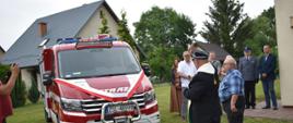 Na zdjęciu widoczny ksiądz święcący nowy samochód ratowniczo gaśniczy przybrany w szarfę biało czerwoną. W tle domek, wokół samochodu zaproszeni goście oraz strażacy osp.
