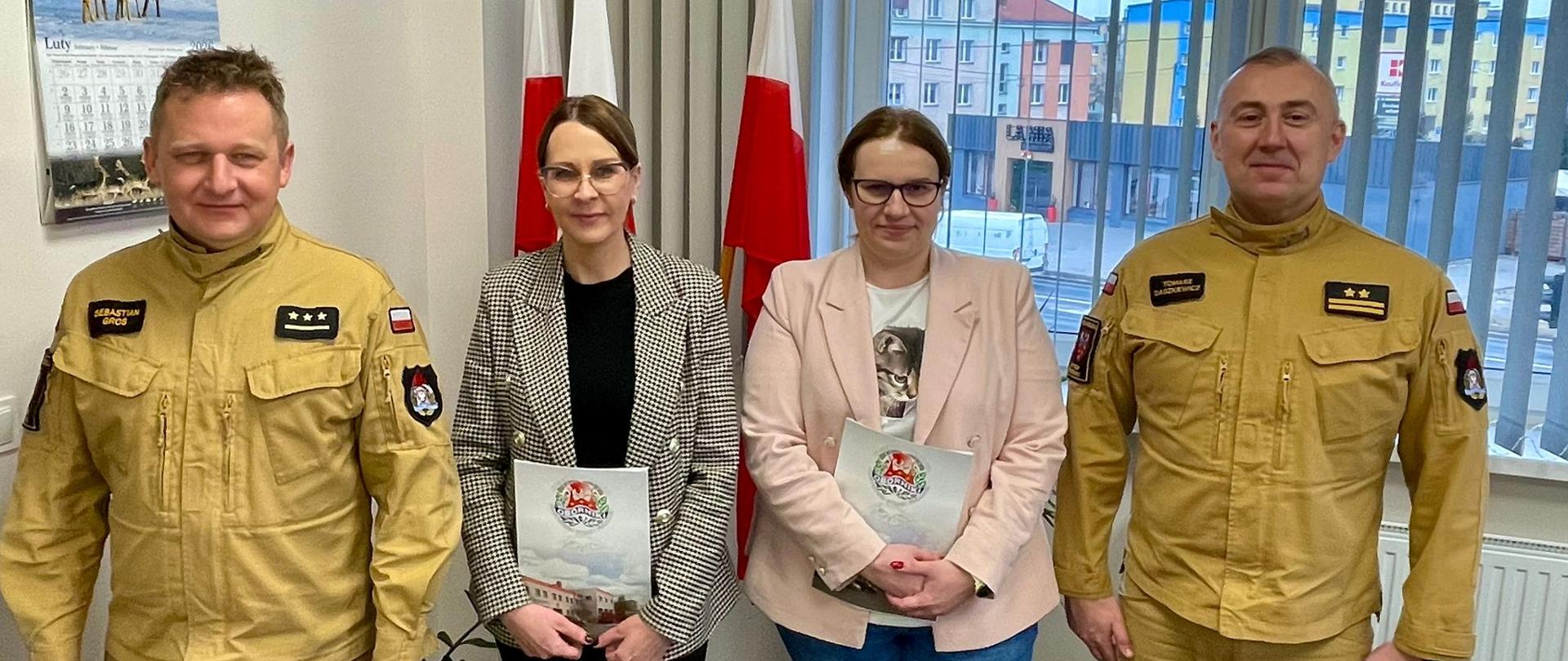 Cztery osoby stoją w biurze na tle trzech flag . Po bokach stoją dwaj mężczyźni w beżowych mundurach służbowych z naszywkami i flagą Polski na ramieniu. Pomiędzy nimi stoją dwie kobiety w strojach cywilnych – jedna w marynarce w kratę, druga w jasnoróżowej marynarce i okularach – trzymające teczki z herbem. W tle widać okno z widokiem na budynki mieszkalne, kalendarz z fotografią jeleni oraz rośliny doniczkowe.