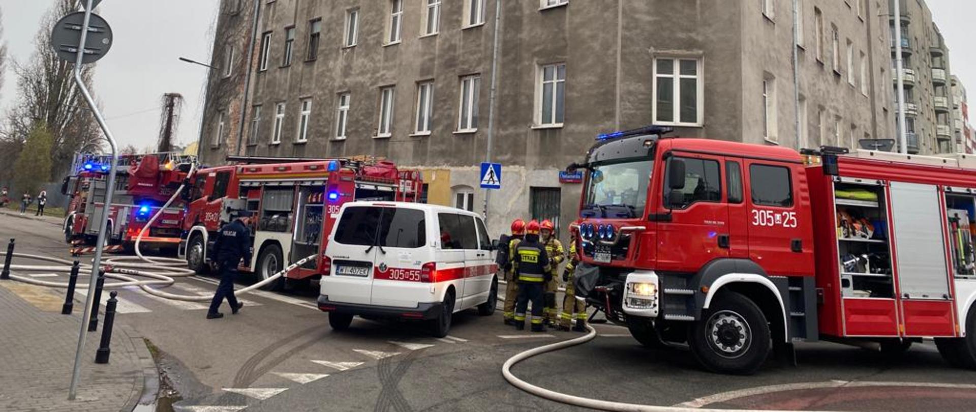Dwa samochody gaśnicze, Samochód specjalistyczny z drabiną mechaniczną oraz biały mikrobus strażaków z Pragi na tle budynku w którym pali się poddasze.