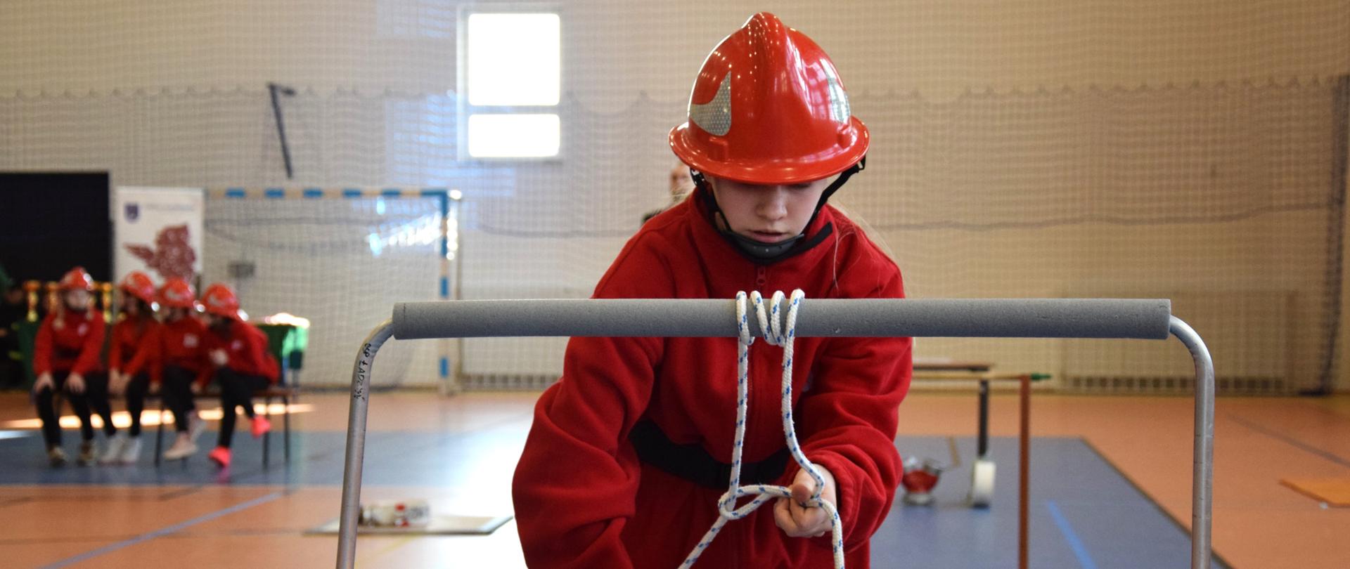 Członek Młodzieżowej Drużyny Pożarniczej podczas pokonywania toru na zawodach sportowych. Dziewczyna zawiązuje węzeł na torze podczas zawodów