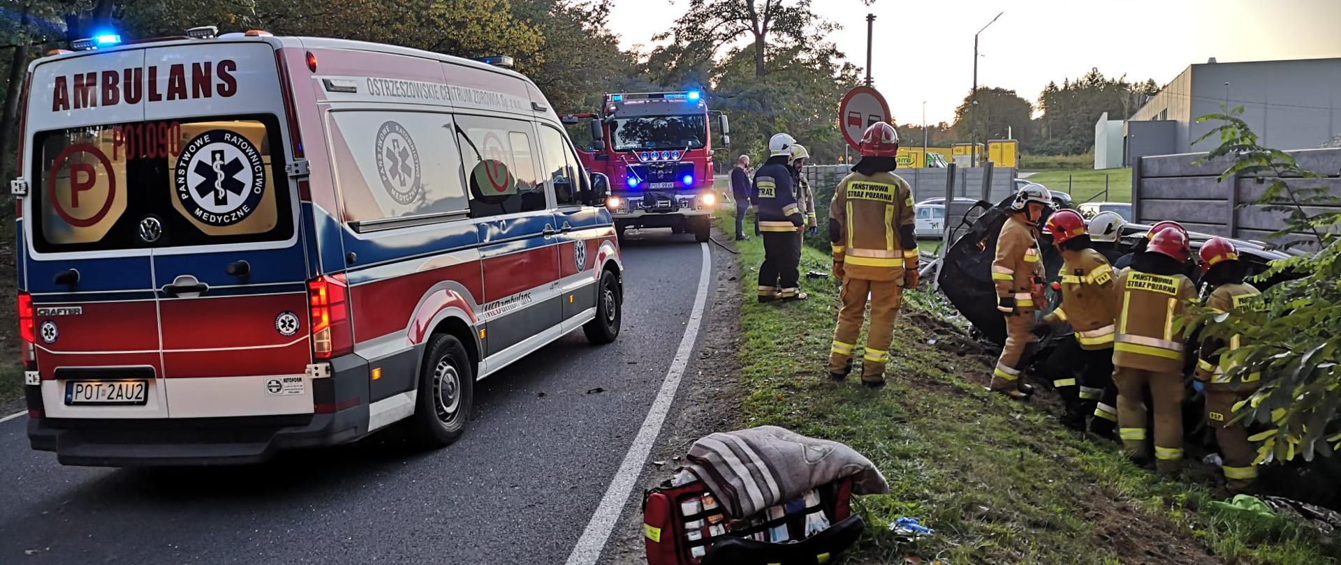 Karetka i wóz strażacki stoją na jezdni, po prawej stronie grupa strażaków znajduje się przy rozbitym pojeździe.